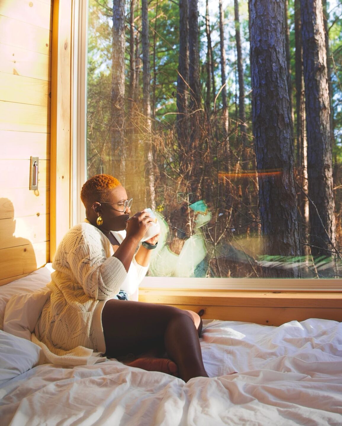 Woman sits on bed by big window looking into nature sipping a cup of coffee.